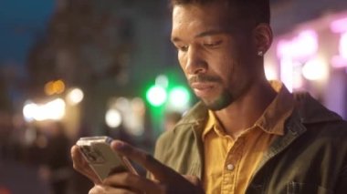 Close-up portrait of focused African American man standing in city center with beautiful neon signs in evening. Busy serious young male tourist chatting online with smartphone. Communication concept.