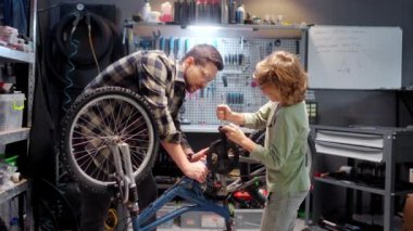 Beautiful caring skillful dad teaching cute little boy to fix bike in authentic workshop with instruments. Attractive caucasian son using tools to repair bicycle with young adult father. Parenting.