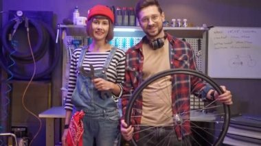 Portrait of young adult handsome skillful craftsman standing with cute beautiful woman mechanic looking at camera smiling holding wheel and tools. Lovely couple fixing bikes in modern garage.