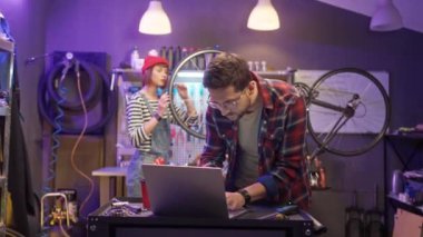 Beautiful caucasian young man using laptop online shopping for bike parts and tools with cute attractive woman fixing bike at modern cycling shop workshop. Lovely couple having business.