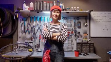 Portrait of beautiful cute happy female mechanic at bike workshop looking at camera smiling standing with crossed arms. Attractive professional woman worker posing at work having good mood.