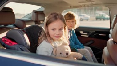 Portrait of cute little blonde girl sitting in back seat of car with beautiful brother looking at camera smiling holding teddy bear. Two lovely kids fastened with safety belts during car trip.