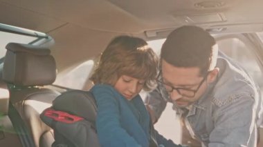 Cheerful attractive loving responsible father fastening cute small son with safety belts preparing before family journey. Lovely happy kid sitting in baby car seat looking forward to car trip.