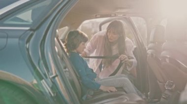 Pretty young caring responsible mother fastening blonde cute little son with safety belts. Happy cheerful boy sitting in back seat of car before family trip bumping fists with father smiling.
