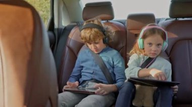 Little cute girl sitting on back seat of car during family car trip with blonde lovely older brother playing games on tablets wearing headphones and listening to music. Safety belts concept.