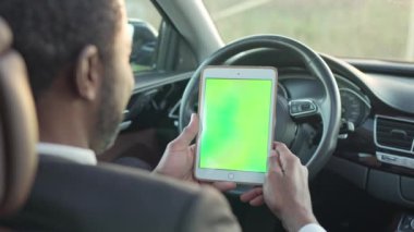 Over-the-shoulder view of attractive wealthy African American man in suit sitting in car holding tablet with green screen. Rich African male businessman using device with chroma key.