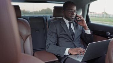 Başarılı bir Afro-Amerikan iş adamının portresi. Arabayla seyahat ediyor, telefonla konuşuyor ve dizüstü bilgisayarında daktilo yazıyor. Takım elbiseli çekici Afrikalı adam iş seyahati yapıyor..
