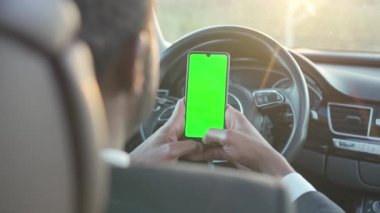 Over-the-shoulder view of influential rich African American man in suit sitting in car holding smartphone with green screen. Successful African businessman holding phone with chroma key.