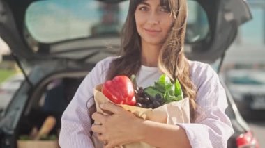 Arabanın yanında duran kızın yakın çekimi. Genç bayan gülümser ve kameraya bakar. Esmer, paketleri poşetlere koyup poz veriyor. Kızın elinde sebze torbası var. Sağlıklı beslenme.