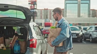 Yakışıklı adam bakkaliye poşetleri taşıyor. Erkek alışverişe çıktı ve arabaya gidiyor. Şık bir adam arabaya alışveriş yapıyor. Sakallı adam paketlerle dolu bagajı kapatıyor. Sebzeli kağıt torbalar..