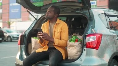 Afro-Amerikalı bir adam arabanın bagajında oturuyor. Ürünlerle birlikte ekolojik paketlerin arka planında bir erkek. Elinde alet ve mesaj tutan çok kültürlü bir adam. Tarz sahibi adam iletişim ve gülümsemeyle tatmin olur..