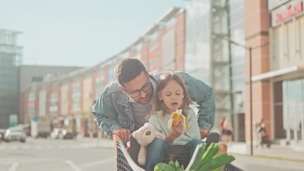 爸爸靠在女儿身边 耳语着 人类购买食物 并把女儿推上购物车 女孩在吃香蕉 阳光普照的父女画像 — 图库视频影像