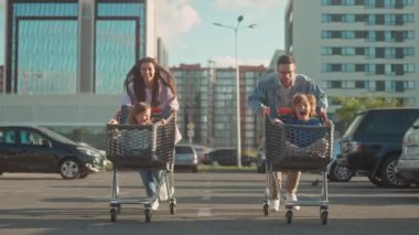 Aile otoparkta yarış oynar. Gülümseyen ebeveynler koşar ve çocukları taşır. Anne ve baba, oğlunu ve kızını market arabalarına itiyor. Mutlu aile boş zamanlarını aktif olarak geçirir. Hafta sonları eğlence..