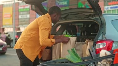 Afro-Amerikalı bir adam satın aldı. Adam alışveriş arabasından bagaja mal taşıyor. Çok kültürlü bir adam eco kağıtlarından alışveriş yapmış. Alışveriş merkezinin arka planındaki müşteri. Etnik çeşitlilik.