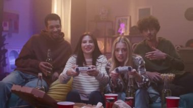 Gençler kanepede oturup video oyunu oynuyorlar. Arkadaşlar eğleniyor ve abur cubur yiyorlar. Gülümseyen ve el kol hareketi yapan insanlardan oluşan bir topluluk. Kız oğlana beşlik çakıyor. Gençliğin akşam eğlencesi.