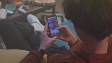 Genç adam sırtından vuruldu. Adam kanepede oturuyor ve telefonu tutuyor. Erkek ekrana dokunuyor ve alete bakıyor. Çocuk odaklanmış ve cihazla çalışıyor. Meslek çeşitliliği.