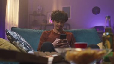 Evin arka planındaki adam. Genç adam kanepede oturuyor ve elinde aletler var. Adam ekrana bakıyor ve gülümsüyor. Kıvırcık saçlı adam konuşarak tatmin olur. Gençlerin internet iletişimi