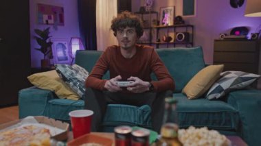 Ön manzara çekimi. Kıvırcık saçlı adam otobüste oturuyor. Genç adam elinde joystick tutuyor ve video oyunları oynuyor. Modern oda ve aperatiflerin arka planında boş vakitleri olan genç yetişkinler..