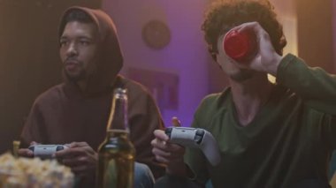 Yavaş çekim. Video oyunu oynayan arkadaşlarının portresi. Afro-Amerikalı adam joystick çiğneyip tutuyor. Genç ayı yataklı adam soda içiyor. İki genç yetişkin iletişim kuruyor ve eğleniyorlar..