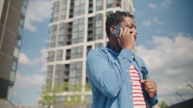 Mavi gömlekli yakışıklı Afro-Amerikan adam telefonundan biriyle konuşuyor. Sokakta dikilirken uzaktan konuşma yapan meşgul bir adam. Açık havada. Teknoloji kavramı.