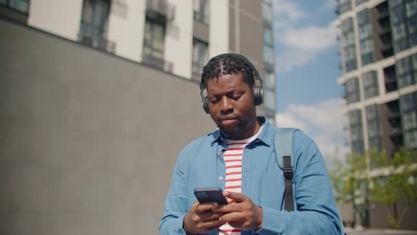 戴耳机的年轻的非裔美国人一边听音乐 一边在手机上输入信息 在街上使用个人技术设备时 认真地看着相机 生活方式 — 图库视频影像