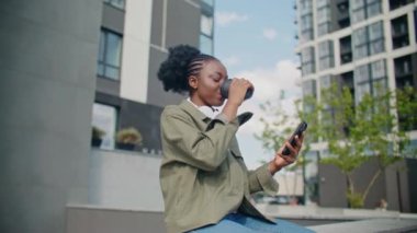 Sokakta sıcak kahve içen Afro-Amerikan bir kadın. Genç bir kadın betonun üzerinde otururken cep telefonunu kullanıyor. Yürürken neşeyle gülümsüyor. Eğlence konsepti.