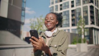 Güneşli bir günde sokakta yürürken kulaklık takan mutlu Afrikalı Amerikalı kadın. Akıllı telefonunu kullanırken neşeyle gülümsüyor. Cihazında birine mesaj yazıyormuş. Teknoloji kavramı.