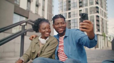 Afro-Amerikalı gençler sokakta eğleniyor. Merdivenlerde oturup mobil cihazın yardımıyla selfie çekmek. Birlikte vakit geçirirken gülmek. Şirin bir çift fotoğraf çekiyor..