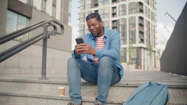 幸せなアフリカ系アメリカ人の男性が階段に座り 誰かにメッセージを入力します 携帯電話を使って笑っている 彼のテクノロジーデバイスで新鮮な空気に時間を費やす — ストック動画