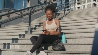 Dışarıda merdivenlerde oturan güzel Afro-Amerikan kadının kamera görüntüsü. Akıllı telefonundan komik videolar izliyor. Su şişesi ve sırt çantası yanında duruyordu. Arka planda koşan bir erkek