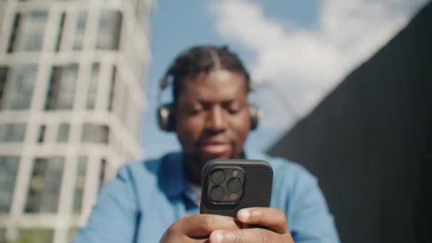 3台のカメラでブラックフォンに集中 魅力的なアフリカ系アメリカ人男性が携帯電話を持ちながらヘッドフォンを着用しています 音楽を聴きながらガジェットを使用しています アーバンストリートシティ — ストック動画