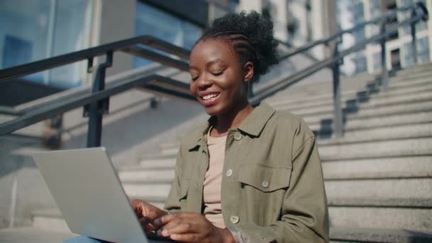 漂亮的非洲裔美国女人穿着衬衫在户外用她的无线手提电脑工作 在和她的伴侣发短信的时候有好感 坐在写字楼附近的楼梯上 — 图库视频影像