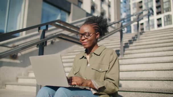 階段に座っている間 ノートパソコンを使ってアフリカ系アメリカ人の学生を魅了する 屋外で試験する準備をしているメガネを持つ若い女性 インターネット上の情報を積極的に閲覧します オンライン教育 — ストック動画