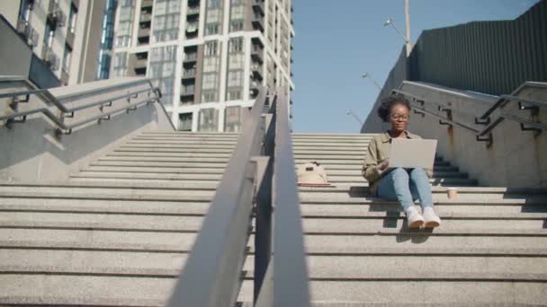 忙碌的非洲裔美国女孩 戴着眼镜 使用她的笔记本电脑 滚动或浏览网站上的东西 晴天坐在现代城市的楼梯上 与互联网的连接 — 图库视频影像