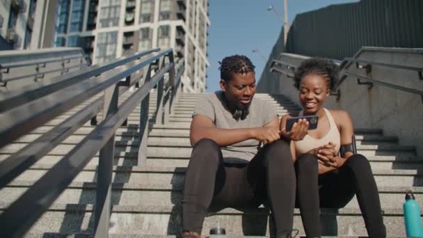 街のどこかでコンクリートの階段に座っている人たち 2人のアフリカ系アメリカ人の友人が一緒に楽しい時間を過ごしています 男性がモバイルデバイスで女性にビデオを見せている 運動後に休憩する — ストック動画