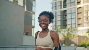 Kamera, dışarıda bekleyen ve doğrudan gülümseyen güzel Afrikalı Amerikalı kadına yaklaşıyor. Şehrin ortasında duran bir kız. Cep telefonu bilekliği ve kremalı spor bluzu giyiyor..