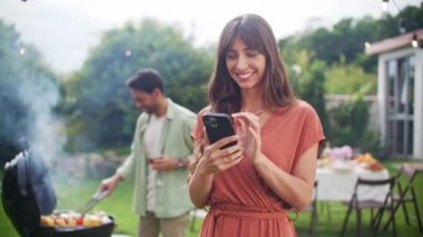 Akıllı telefonu tutan, gülümseyen, mesajlaşan, sohbet eden ve telefon ekranına bakan güzel bir kadının portresi. Tatil, tatil, yemek, insanlar ve yemek konsepti - arkadaşlar ev bahçesinde parti veriyor.