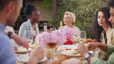 Kutlama, arkadaşlık ve tatil konsepti. Çok ırklı genç kadın ve erkeklerden oluşan bir grup evin arka bahçesindeki masada oturmuş yemek yiyor, iletişim kuruyor ve şampanya bardaklarını tokuşturuyorlar..