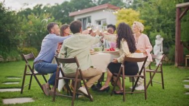 Çok ırklı genç erkek ve kadınlardan oluşan bir grup evin arka bahçesindeki masada oturmuş yemek yiyor, iletişim kuruyor ve şampanya kadehlerini tokuşturuyorlar. Kutlama, arkadaşlık ve tatil kavramı.