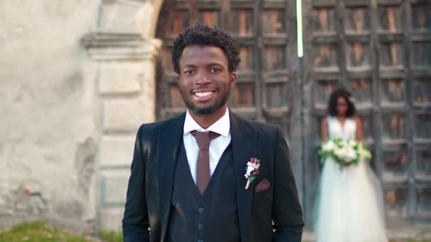 エレガントなスーツを着たアフリカ系アメリカ人男性は カメラで直接微笑んでいます 幸せな結婚式の日を楽しむ魅力的な花婿 バックグラウンドスタンディングフィアンセと花の大きな花束 — ストック動画