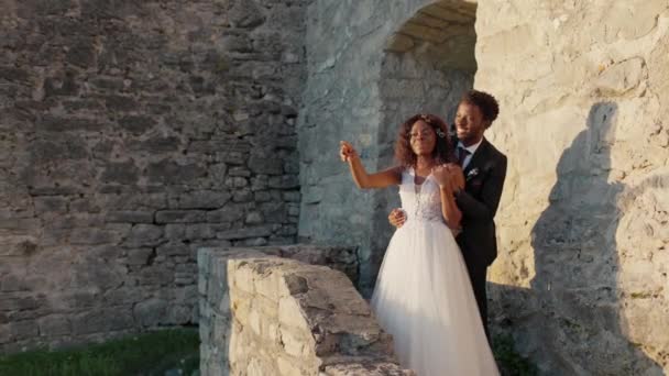 身穿婚纱的非洲裔美国夫妇站在石头城堡里看着日落 年轻的丈夫拥抱他可爱的妻子 一起笑着 愉快的气氛 爱和庆祝的概念 — 图库视频影像