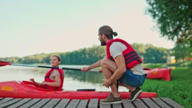 Kızıl yelek giyen beyaz sakallı bir adam, kadınına kanoyla yardım ediyor. Sakallı erkek, göl kenarında otururken kameraya neşeyle gülümsüyor. Çiftler yaz tatilinde doğada..