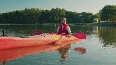 Çekici beyaz bir adam kanoda oturuyor. Gölde dinlenirken kırmızı güvenlik yeleği giyiyor. Aksiyon kameralı bir tekne. Güneşli bir günde kano gezisi. Sucul sporları. Yakışıklı adam doğada eğleniyor..