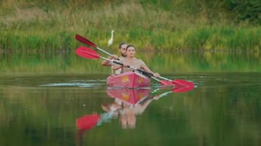 Kafkasyalı çiftin kameraya doğru kürek çekerken ön görüntüsü. Arka planda beyaz balıkçıl. Sevgili karı koca sinerjik bir şekilde büyük gölün ortasında kürek çekiyorlar. Aktif olarak hafta sonu geçirmek.