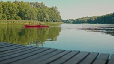 Kırmızı yelekli bir kadın büyük göl boyunca kanoyla kürek çekiyor. Teknede otururken küreği tutmak. Açık hava sporlarını seviyorum. Güneşli bir günde kano yaparken eğleniyorum. Yaşam tarzı. Doğada yolculuk.