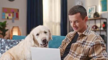 Çekici beyaz adam rahat odasında köpeğiyle birlikte kanepede oturuyor. Laptop kullanan ve altın labrador av köpeğini okşayan pozitif bir erkek. Hafta sonu evde dinleniyorum. Hayvan konsepti.