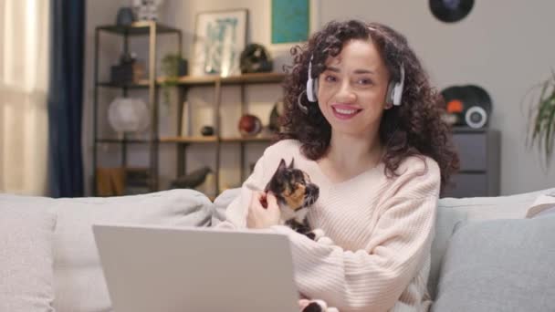 可爱的白人女人 带着耳机和笔记本电脑坐在家里柔软的沙发上 漂亮的工人抱着她的毛茸茸的猫在房间里远程工作 看着相机 开心地笑着 — 图库视频影像