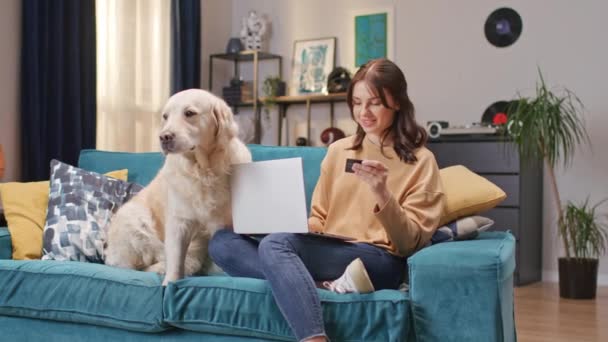 带着笔记本电脑的快乐女人在网上为她可爱的小狗买食物 漂亮的买主用信用卡购物 而她的毛茸茸的狗坐在沙发上 对成功购买感到满意 — 图库视频影像