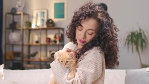 若い白人女性が小さな赤い猫を抱きしめ 喜んでカメラに微笑んでいます 家で居心地の良い部屋で彼女の素敵な子猫を飼っているカーリーの女性の所有者 動物への愛の概念 — ストック動画