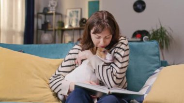 Çekici beyaz kadın hoş Jack Russell teriyeriyle oturma odasında dinleniyor. Şirin köpeğine sarılıp okşayarak kitap okuyan güzel bir bayan. Evde birlikte dinleniyoruz..